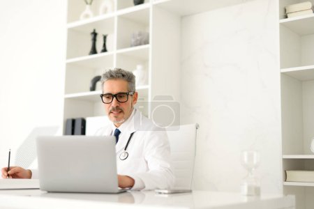 Photo for Mature, grey-haired doctor deeply engrossed in his work on a laptop, possibly consulting a medical database or researching for patient treatment, taking notes with serious expression reflecting - Royalty Free Image