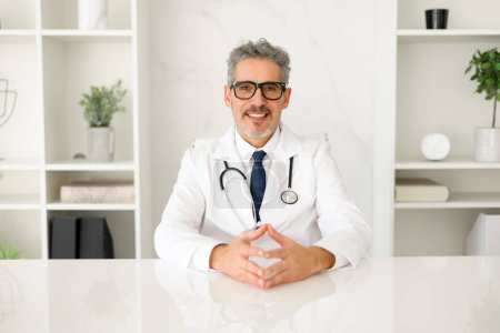Photo for A friendly senior doctor looking directly at the camera, his hands open in a welcoming gesture, inviting trust and communication in a modern healthcare setting. Portrait of experienced MD on workplace - Royalty Free Image