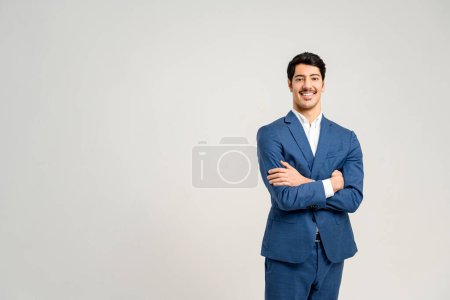 Photo for Cheerful young professional in smart blue suit with crossed arms showcases confidence and a readiness for business challenges, set against clean, light background that accentuates his sharp appearance - Royalty Free Image