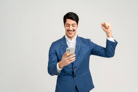 A man in a crisp blue suit beams with success as he celebrates a victory or achievement on his tablet, against a simple background that keeps the focus on him.