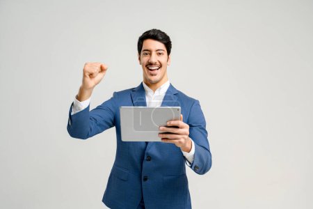Téléchargez les photos : Un homme d'affaires joyeux en costume bleu montre un geste triomphant en regardant une tablette, symbolisant une réussite numérique réussie ou une percée, isolé sur blanc - en image libre de droit