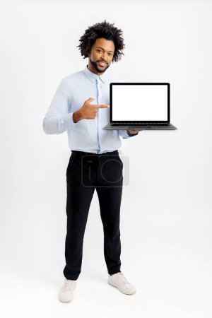 Téléchargez les photos : Homme d'affaires afro-américain souriant en tenue formelle montrant ordinateur portable avec affichage vide isolé sur blanc. Employé masculin pointe du doigt à l'écran blanc, la publicité nouvelle application, présentation de newelty - en image libre de droit