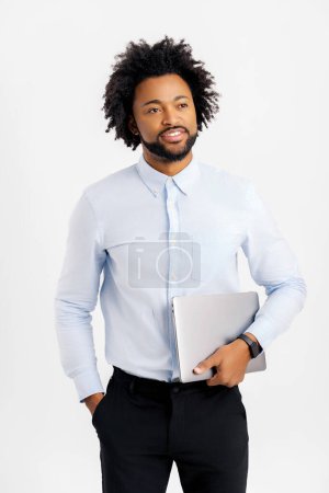 Téléchargez les photos : Un jeune entrepreneur afro-américain souriant se tient dans un studio isolé sur un ordinateur portable blanc et branché. gai noir guy dans formelle usure tenue fermé ordinateur portable - en image libre de droit
