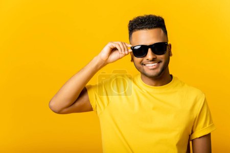Foto de Guapo joven indio atractivo en gafas de sol posando aislado sobre fondo amarillo. Despreocupado joven masher hombre mirando a la cámara y riendo, coqueteando con usted - Imagen libre de derechos