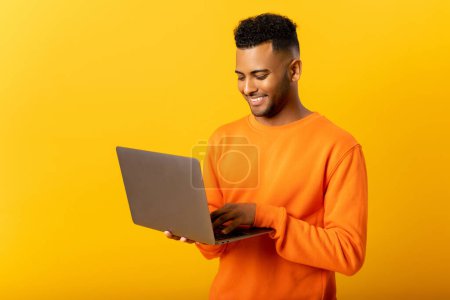 Foto de Retrato de un joven indio de pie aislado en una computadora portátil amarilla, escribiendo mensajes, charlando con un amigo en las redes sociales en línea, viendo video, disfrutando de una nueva aplicación, websurf - Imagen libre de derechos