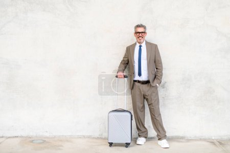 Photo for With a bright smile and a casual stance, a senior experienced businessman stands with his suitcase against concrete wall, embodying the ease and comfort of contemporary business travel - Royalty Free Image