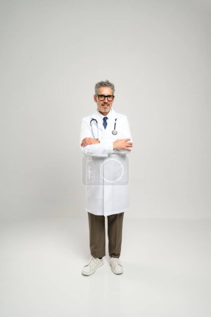 Ein Oberarzt in weißem Mantel und Brille, selbstbewusst mit verschränkten Armen vor einem schlichten Hintergrund stehend, spiegelt sein erfahrenes Fachwissen und das Vertrauen wider, das er seinen Patienten einflößt..