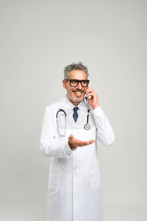 Ein gut gelaunter Oberarzt mit grauen Haaren spricht auf einem Smartphone, gestikuliert, als würde er ein Konzept erklären oder Ratschläge erteilen, vor reinem, weißem Hintergrund, in weißem Mantel und Stethoskop.