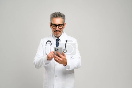 Un médico senior con un comportamiento amigable está sosteniendo un teléfono inteligente, revisando la información médica o programando citas, con un estetoscopio alrededor de su cuello y una mirada de concentración.