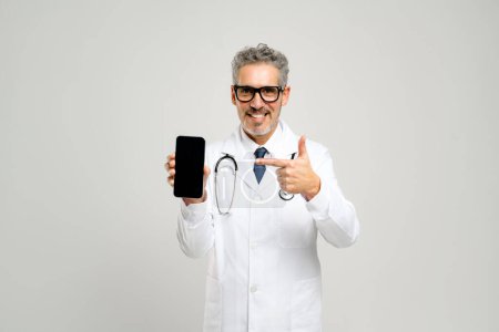 Ein reifer Arzt geht mit dem Publikum ins Gespräch und zeigt auf einen leeren Smartphone-Bildschirm, auf dem medizinische Daten oder eine Gesundheitsanwendung angezeigt werden können, im weißen Kittel isoliert stehend