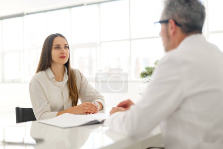 Photo for A senior doctor in a white coat engaging with a patient in a conversation that appears both professional and warm, medical office provides a clean and calm backdrop for the consultation - Royalty Free Image