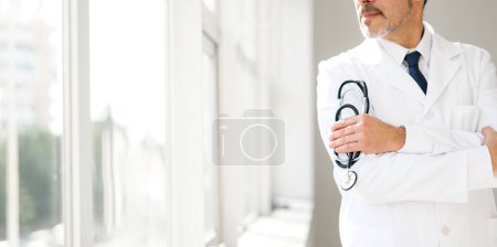 Ein nachdenklicher Oberarzt mit Stethoskop, der im weißen Mantel am Fenster steht und in seiner Haltung Erfahrung und Kontemplation vermittelt. Breites Banner, abgeschnittene Ansicht, Gesicht nicht sichtbar