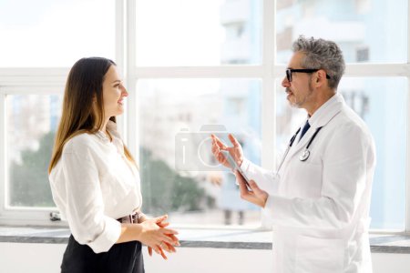 Photo for A senior doctor engaged in a conversation with a young female patient, both looking at a tablet in a sunlit, modern medical office. The picture reflects a commitment to patient-centered care - Royalty Free Image