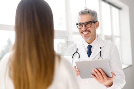 Ein angesehener Arzt mit grauen Haaren hält eine Tablette in der Hand und unterhält sich mit einer jungen Patientin in einer hellen, modernen Klinik.