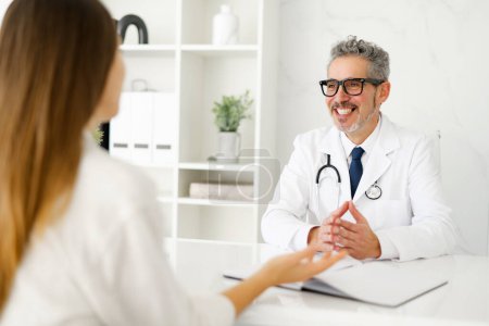 A senior male doctor with grey hair and glasses is seen smiling reassuringly at a female patient, creating a warm and welcoming atmosphere in a modern, well-lit consultation room.
