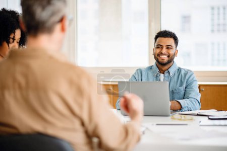 A young Latin man in casual wear, with a beaming smile participates in a meeting, his attentiveness and joy reflecting the positive dynamics of a diverse and modern office team.
