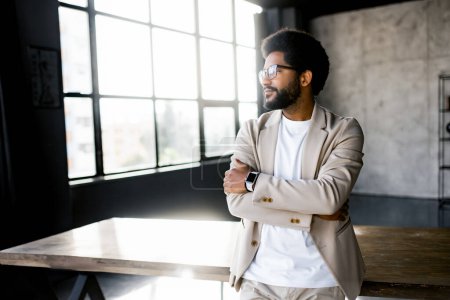 Photo for A confident young businessman stands with arms crossed in a sunlit modern office, gazing thoughtfully out of large windows, embodying success and professionalism - Royalty Free Image