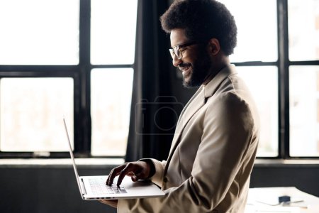 The young professional stands by the window, his posture relaxed as he uses his laptop, bathed in the natural light that emphasizes a productive day at the office. The large windows in the background