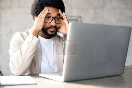 Photo for A young businessman appears troubled, holding his head while staring at a laptop, a representation of work stress or problem-solving in a professional setting - Royalty Free Image
