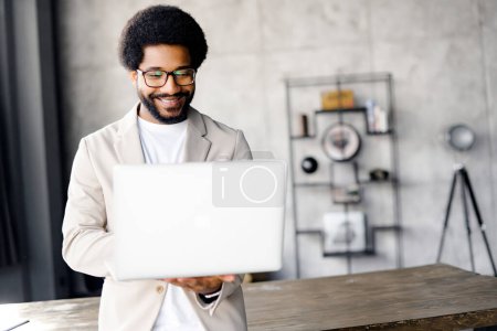 A young, stylish businessman in a beige blazer stands holding a laptop, his face expressing concentration and professionalism. The modern industrial-style office setting adds to the contemporary vibe