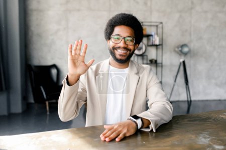 A cheerful young businessman greets with waving during virtual meeting, seated in a modern office environment, exuding a vibe of friendly professionalism. Virtual meeting concept