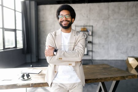 Téléchargez les photos : Un homme d'affaires afro-américain joyeux avec un sourire radieux se tient avec confiance dans un environnement de bureau spacieux, sa montre intelligente complétant son look professionnel moderne - en image libre de droit