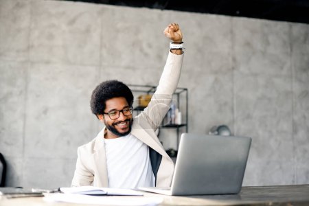 Elation and success radiate from the young businessman as he celebrates an achievement, raising his fist in a triumphant gesture working at his laptop, feels accomplishment and career satisfaction