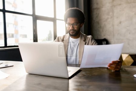 Ein fokussierter Geschäftsmann analysiert ein Dokument neben seinem Laptop in einem hellen, modernen Büroraum und verkörpert die Verschmelzung traditioneller Geschäftspraktiken mit modernen digitalen Werkzeugen