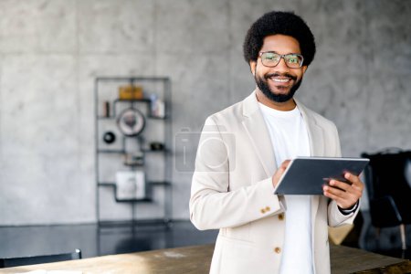 Photo for The young office professional holding a tablet, standing against an industrial backdrop, a modern, creative work environment. His smile suggest a friendly approach to business - Royalty Free Image