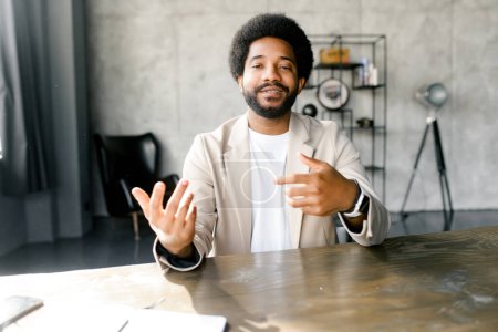 Ein junger brasilianischer Geschäftsmann, der an einem virtuellen Meeting teilnimmt, kommuniziert selbstbewusst, seine Hände übermitteln seine Botschaft in einem modernen Büro, der Mann blickt in die Kamera und spricht