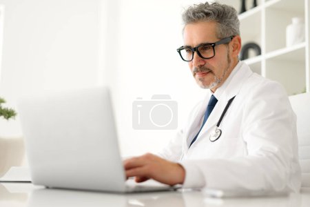 Senior doctor with grey hair and black-rimmed glasses focuses intently on a laptop screen, symbolizing the modern healthcare professionals reliance on technology for patient care and medical research
