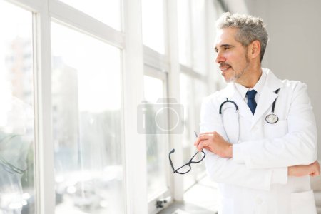 Ein Oberarzt in weißem Mantel steht in einem hellen Klinikzimmer, mit sanftem Gesichtsausdruck und Brille, und reflektiert einen Moment der Atempause oder tiefer Gedanken inmitten eines geschäftigen Tages.