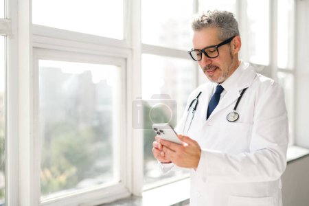 With a confident and friendly smile, this senior doctor engages with his smartphone, navigating medical software or patient communication apps
