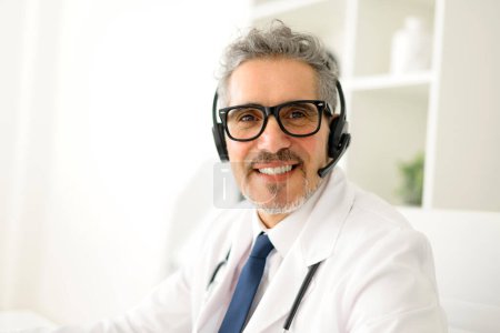 Foto de Un médico de cabello gris equipado con un auricular sonríe calurosamente, listo para proporcionar servicios de telesalud, mostrando el enfoque moderno de la atención al paciente accesible e inmediata - Imagen libre de derechos