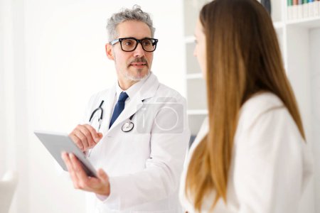 Photo for A senior doctor, with a stethoscope around his neck, attentively listens to the young female patient while holding a tablet, symbolizing a patient-centered approach in a clinical setting - Royalty Free Image