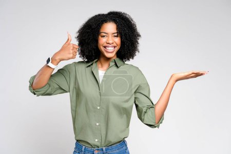 Téléchargez les photos : Une femme afro-américaine exubérante aux cheveux bouclés sourit largement, donnant un pouce levé avec sa main droite tandis que sa main gauche est ouverte comme si elle présentait, recommandait ou approuvait quelque chose - en image libre de droit