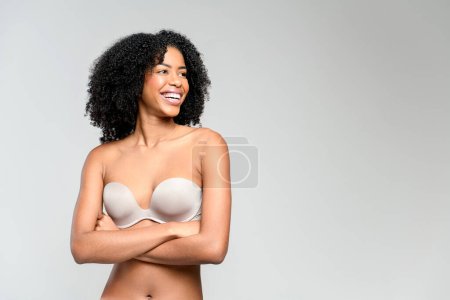 A gleeful African-American woman in a strapless top chuckles heartily with her arms crossed, standing against a soft gray backdrop. Image captures the essence of genuine mirth and relaxed comfort