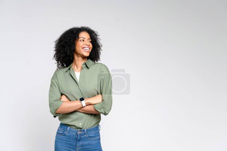 Foto de Una mujer afroamericana segura de sí misma con una camisa verde oliva casual y pantalones vaqueros se levanta con una sonrisa radiante, exudando un profesionalismo relajado y un encanto relajante sobre un fondo claro. - Imagen libre de derechos