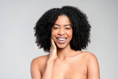 A captivating African-American woman with an infectious laugh and her hand gently caressing her face stands bare-shouldered against a muted background, presenting an image of unadulterated joy