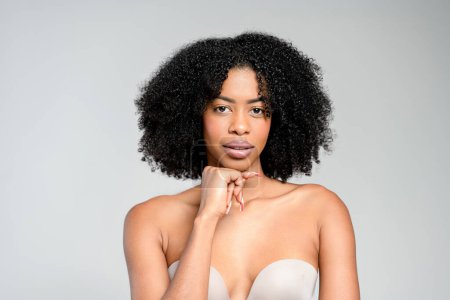An African-American woman with striking natural curls and a thoughtful expression poses in a strapless top, her hand delicately resting on her chin, against a serene gray backdrop