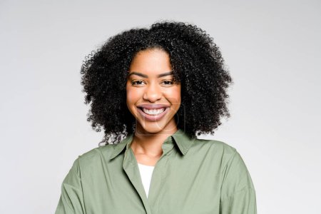 Una mujer afro-americana alegre en una camisa de oliva elegante-casual posa y mira a la cámara, su sonrisa radiante y comportamiento confiado proyectando fuerza y accesibilidad sobre un fondo neutral.