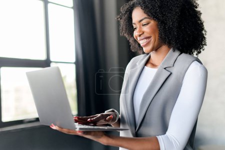Téléchargez les photos : Femme d'affaires afro-américaine utilise un ordinateur portable dans un bureau moderne, son sourire rayonnant et tenue élégante reflétant une image d'entreprise réussie. L'ambitieux professionnel dans un espace de travail productif. - en image libre de droit