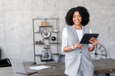 Téléchargez les photos : Une femme d'affaires afro-américaine bien préparée tient une tablette dans son bureau bien aménagé, son sourire éclatant et son élégance décontractée soulignant un mélange de professionnalisme moderne et d'accessibilité - en image libre de droit