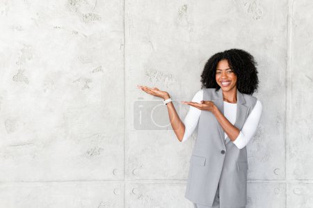 With arms pointing aside, a joyful African-American businesswoman presents an invisible object, her engaging smile and open stance suggesting a friendly invitation to potential opportunities