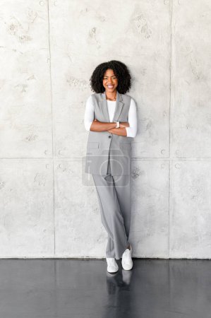 Una mujer de negocios afroamericana profesional se levanta con confianza, con los brazos cruzados, con una pared gris en el fondo, exudando liderazgo y aplomo