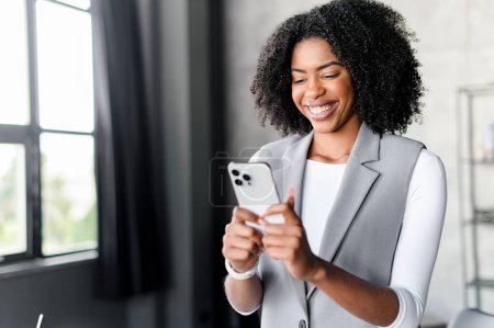 African-American businesswoman is captured in a candid moment of joy, browsing on her smartphone by the window, bathed in natural light that highlights her confident, tech-savvy presence.