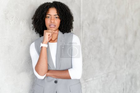 Femme d'affaires afro-américaine en costume gris élégant se tient contemplativement, son regard fixé soigneusement dans la caméra, incarnant le professionnalisme et la pensée avant-gardiste dans un monde d'entreprise moderne