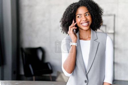 Mit einem strahlenden Lächeln unterhält sich eine afroamerikanische Geschäftsfrau am Telefon und verbindet mühelos professionellen Scharfsinn mit moderner Konnektivität im Büro, effektiver Kommunikation im digitalen Zeitalter