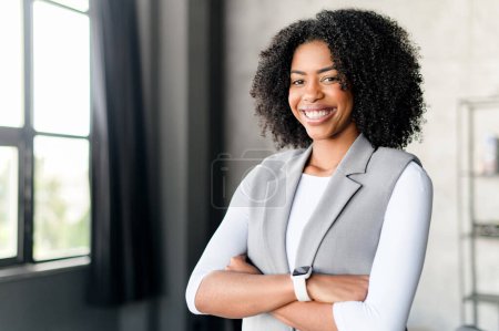 A cheerful African-American businesswoman, clad in a modern grey vest and white sleeves, stands with her arms crossed, showcasing a smile that communicates friendliness and professionalism