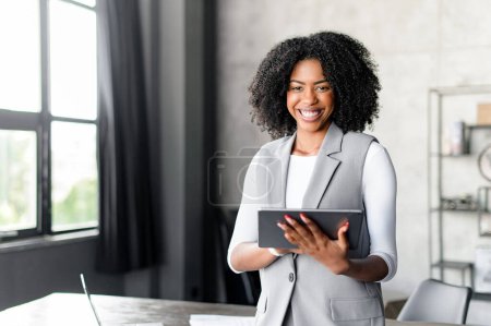 Foto de Una alegre empresaria afroamericana sostiene con confianza una tableta digital en un entorno de oficina moderno, su sonrisa expresando satisfacción y profesionalismo - Imagen libre de derechos